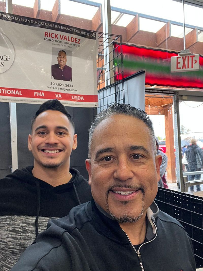 Two men smiling, posing for a selfie in front of a banner for Rick Valdez, at a market or event.