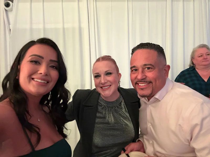Three people smiling for a selfie; woman with dark hair in a green dress, woman in blazer, man in sweater.