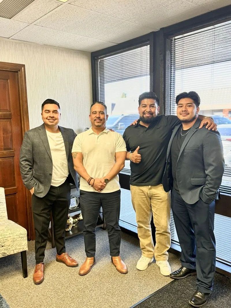 Four men standing indoors. Three wear jackets and dress pants; the fourth, a polo shirt and khakis, gives a thumbs-up.