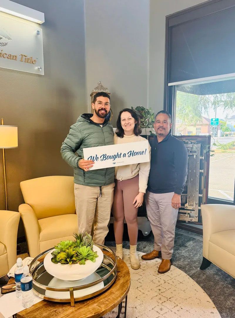 Three people pose indoors, holding a sign. Two chairs flank a coffee table with a plant. Sunlight streams through a window.