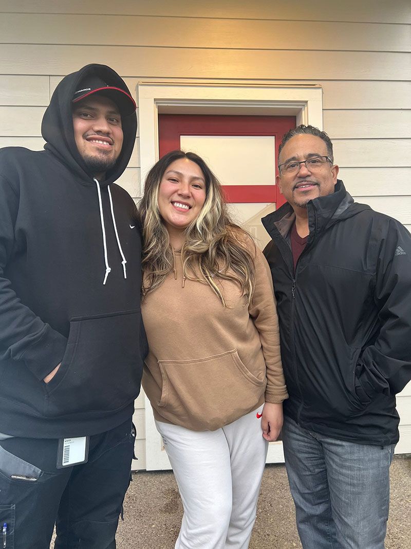 Three people smiling in front of a building with a red door.