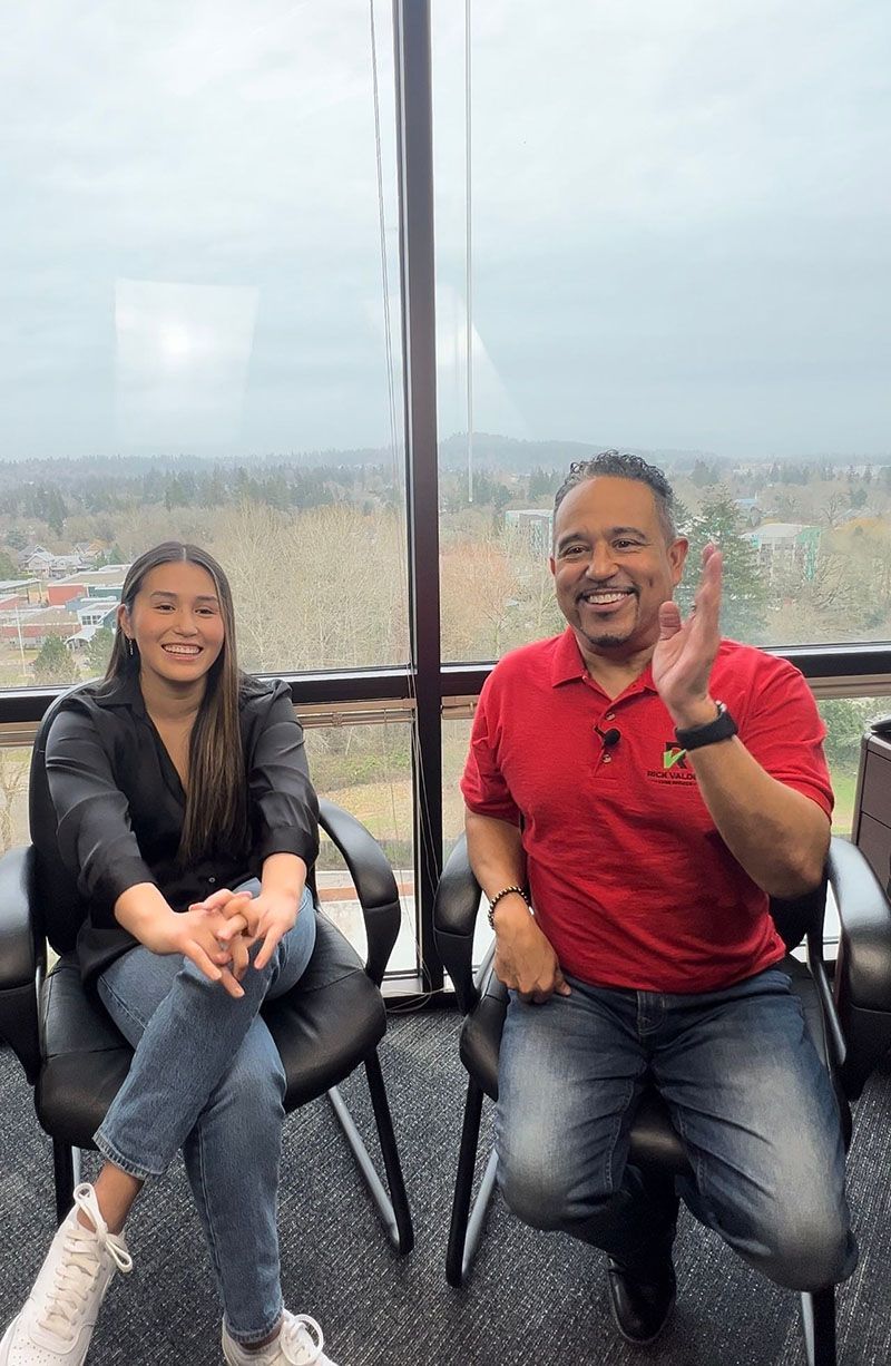 Woman and man seated, smiling, gesturing, by a window overlooking a cityscape; man wears red shirt.