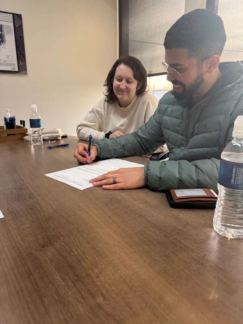 Man in green jacket signs a paper at a table, smiling. Woman watches. Bottles and pen on the table.