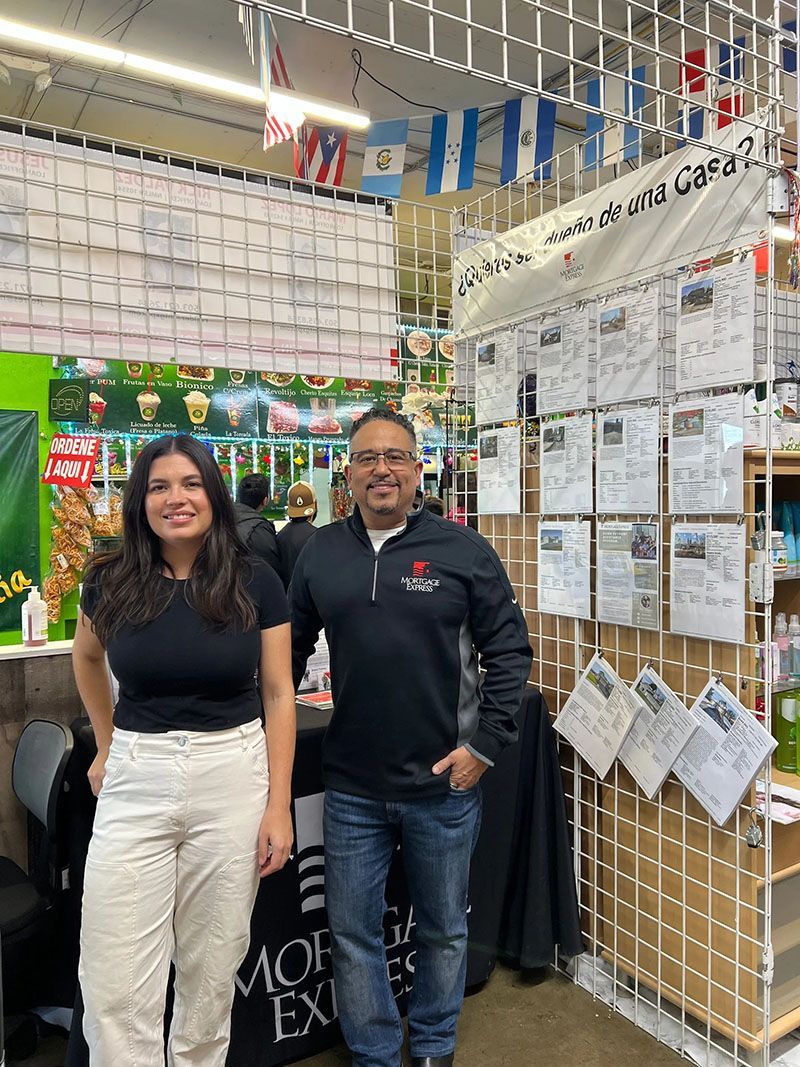 Two people stand in front of a market stall with flags and signs. One woman and one man smile.