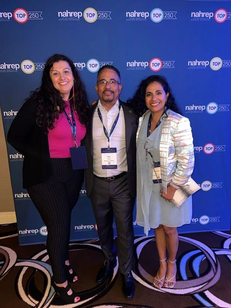 Three people pose in front of a blue backdrop with event logos. One man and two women smile.