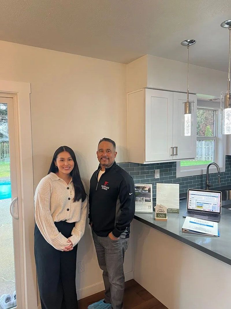 Two people stand in a kitchen; woman in a sweater, man in a jacket. Counter with brochures and laptop.