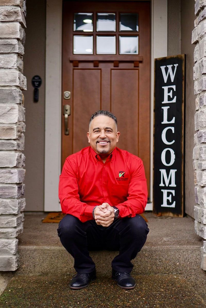 Man in red shirt kneels on porch, hands clasped, in front of a welcome sign and wooden door.