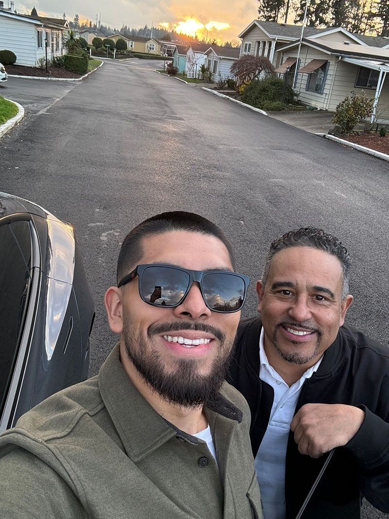Two smiling men take a selfie outside, mobile homes in background, sunset.