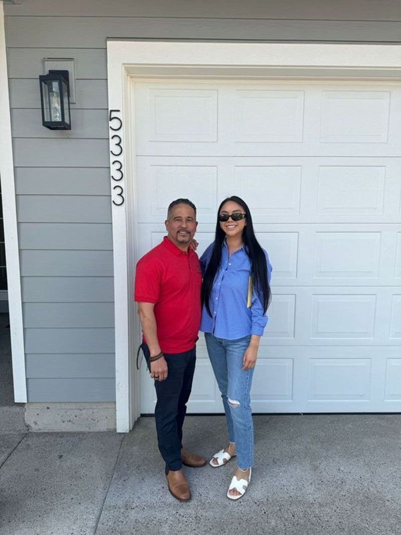 A man and woman stand in front of a white garage door. The man wears a red shirt, the woman a blue shirt.