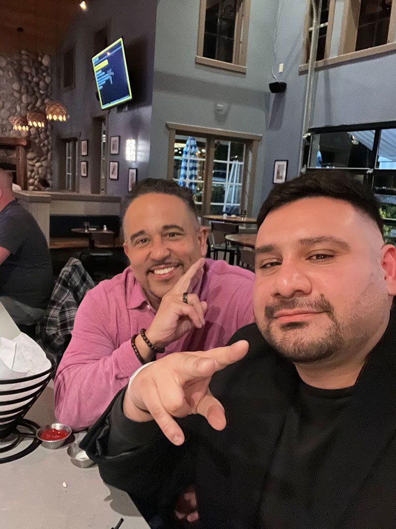 Two men at a restaurant, one points at camera, the other smiles. Pink shirt, black top, interior setting.