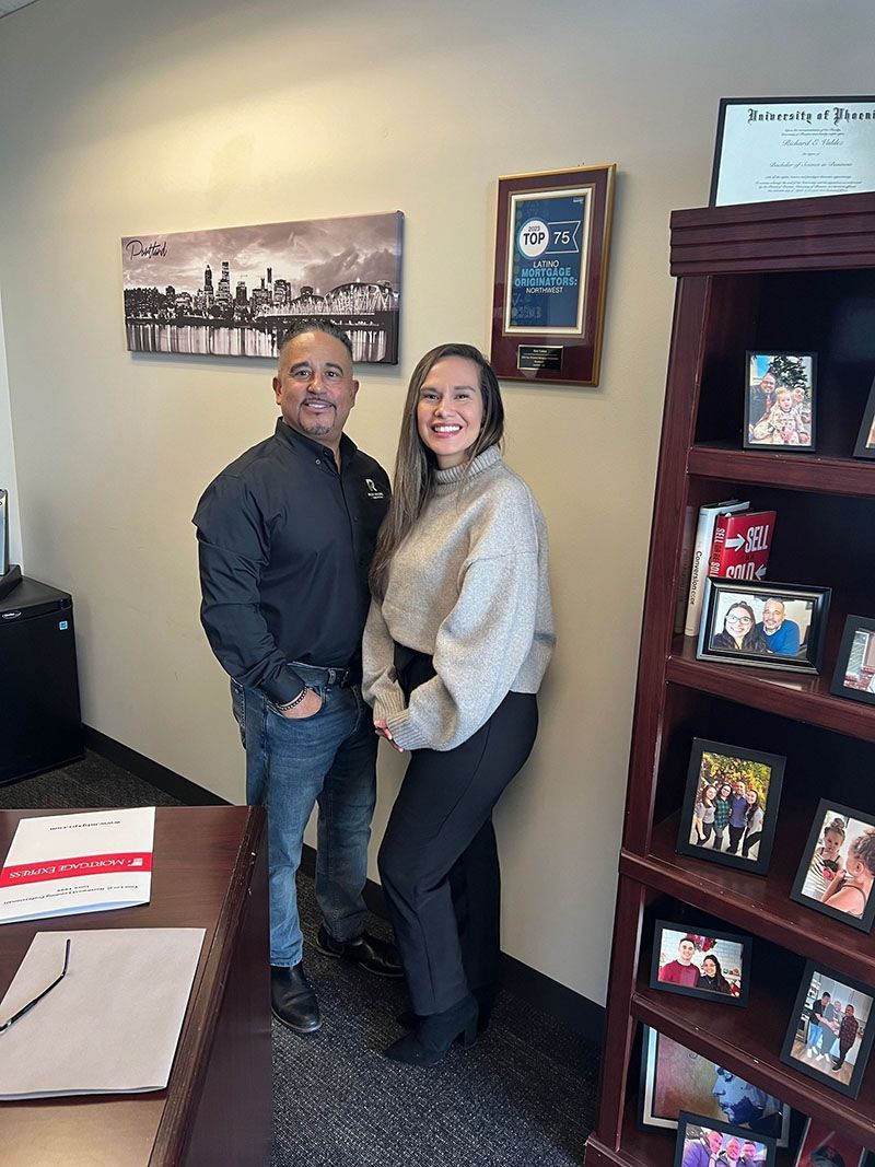 Two people standing in an office with a bookshelf. The man wears a black jacket and jeans. The woman wears a cream sweater and black pants.