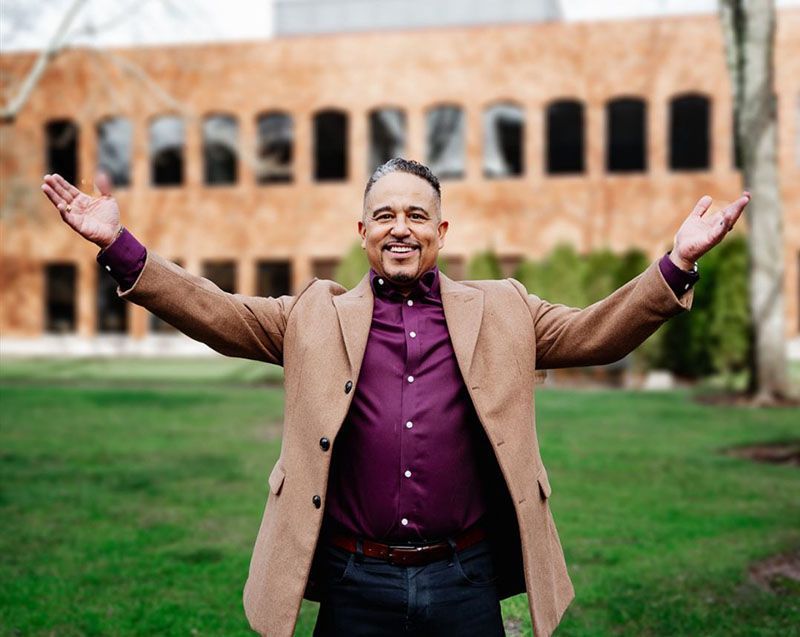Man with arms outstretched, smiles outdoors; wearing a tan blazer, purple shirt, and jeans, in front of a brick building.