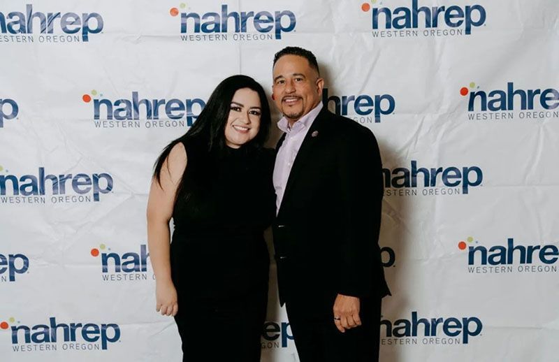 Two people posing in front of a backdrop with logos. Woman in black dress, man in suit.
