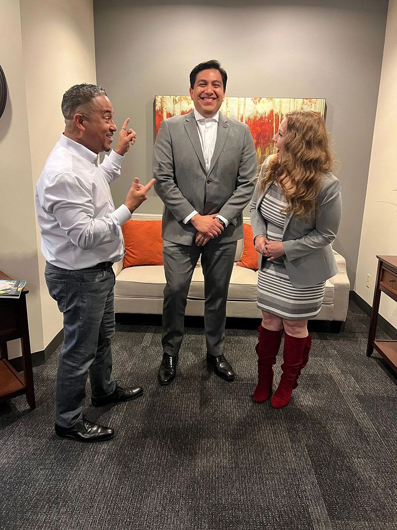 Three people stand in an office: one points, one smiles, and one is beside them. A couch and art are in the background.