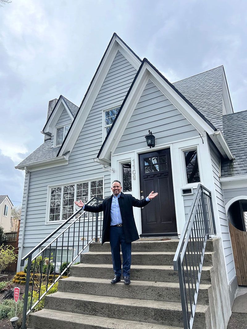 Man standing on steps of a light gray house with arms outstretched. Dark door, white trim, cloudy sky.