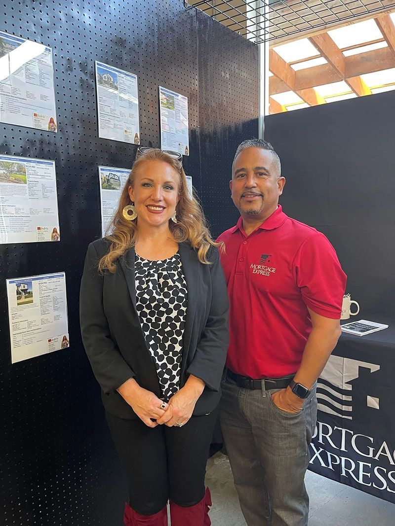 Woman and man standing by display with paperwork. Woman in black blazer, man in red shirt.