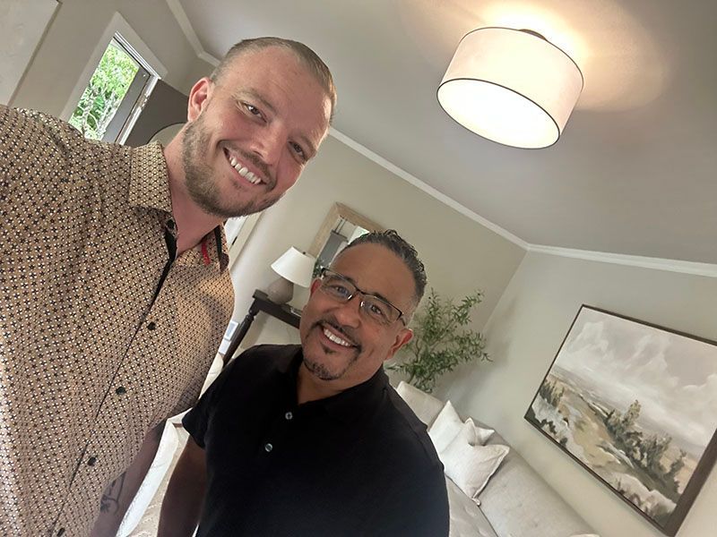 Two men smile for a selfie in a brightly lit room. One wears a patterned shirt, the other a black shirt.