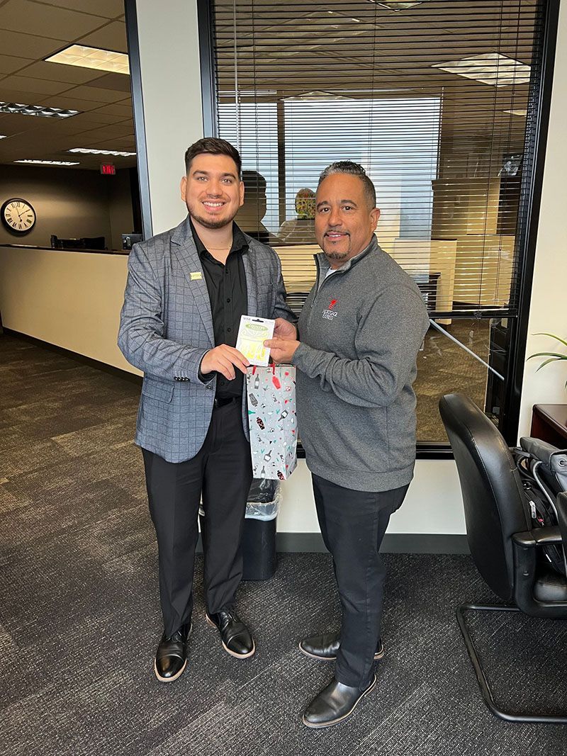 Two men in an office setting, one handing a gift. Man on left wearing a blazer. Man on right in a grey sweatshirt.