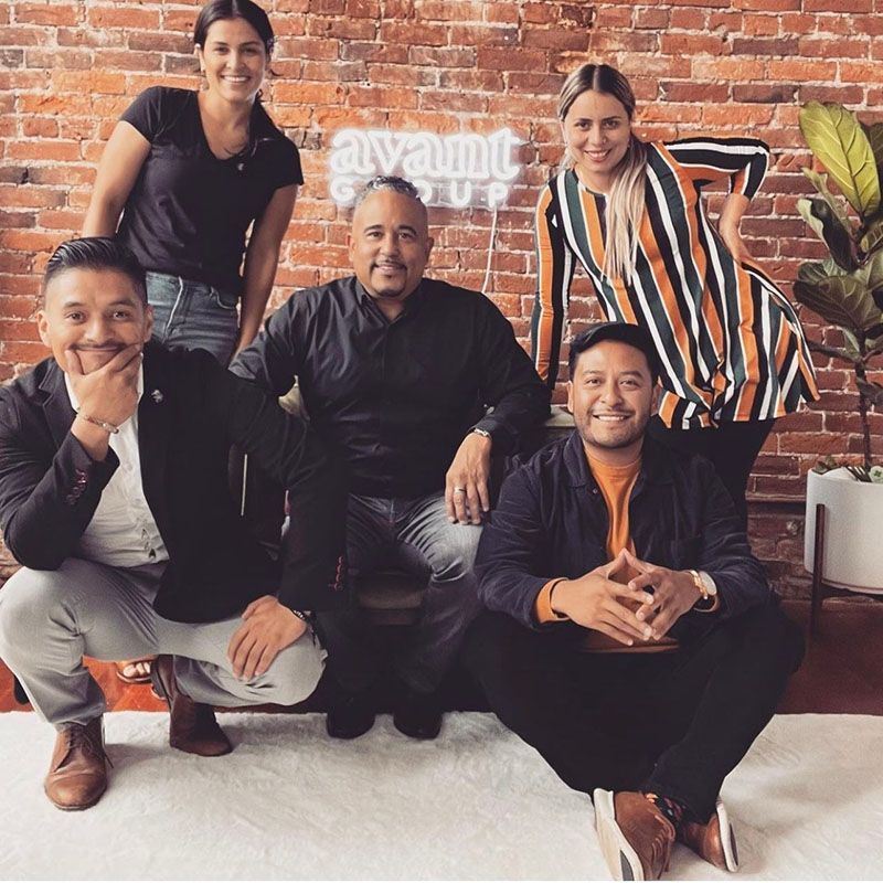 Group of five people posing in front of a brick wall with neon sign 