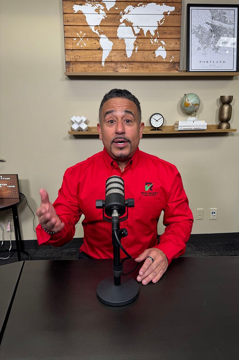 Man in red shirt speaking into a microphone at a desk; a world map and shelf with décor are in the background.