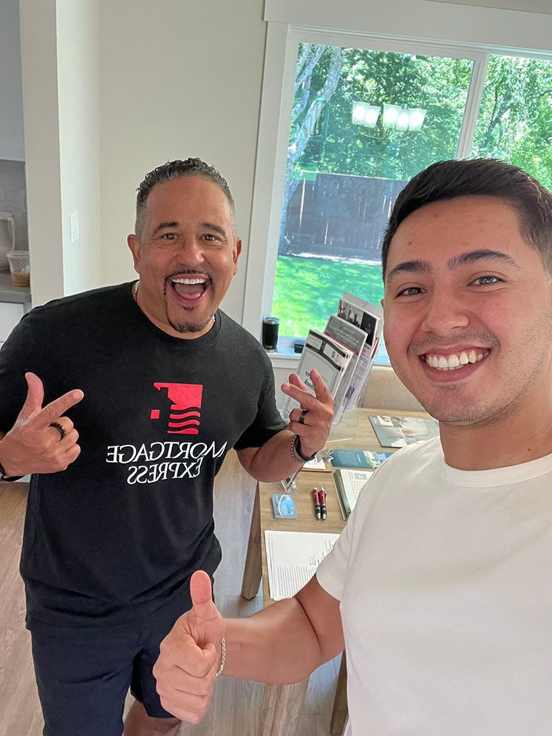 Two men smiling in a room; one points, the other gives a thumbs-up. Sample materials sit on a table.
