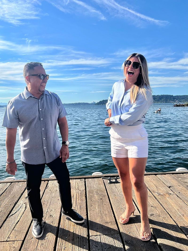 Man and woman laughing on a wooden dock with water and blue sky in background.