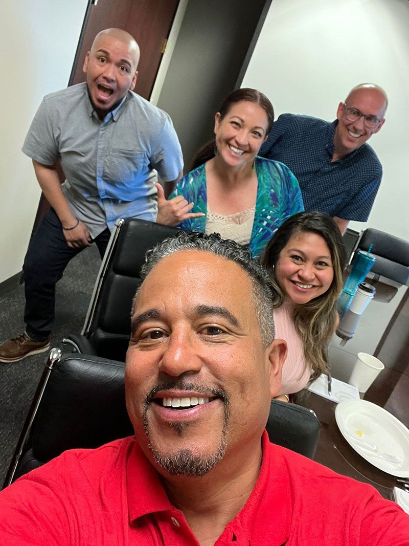 Group of five people smiling, making expressions, in an office. One takes a selfie in the foreground.