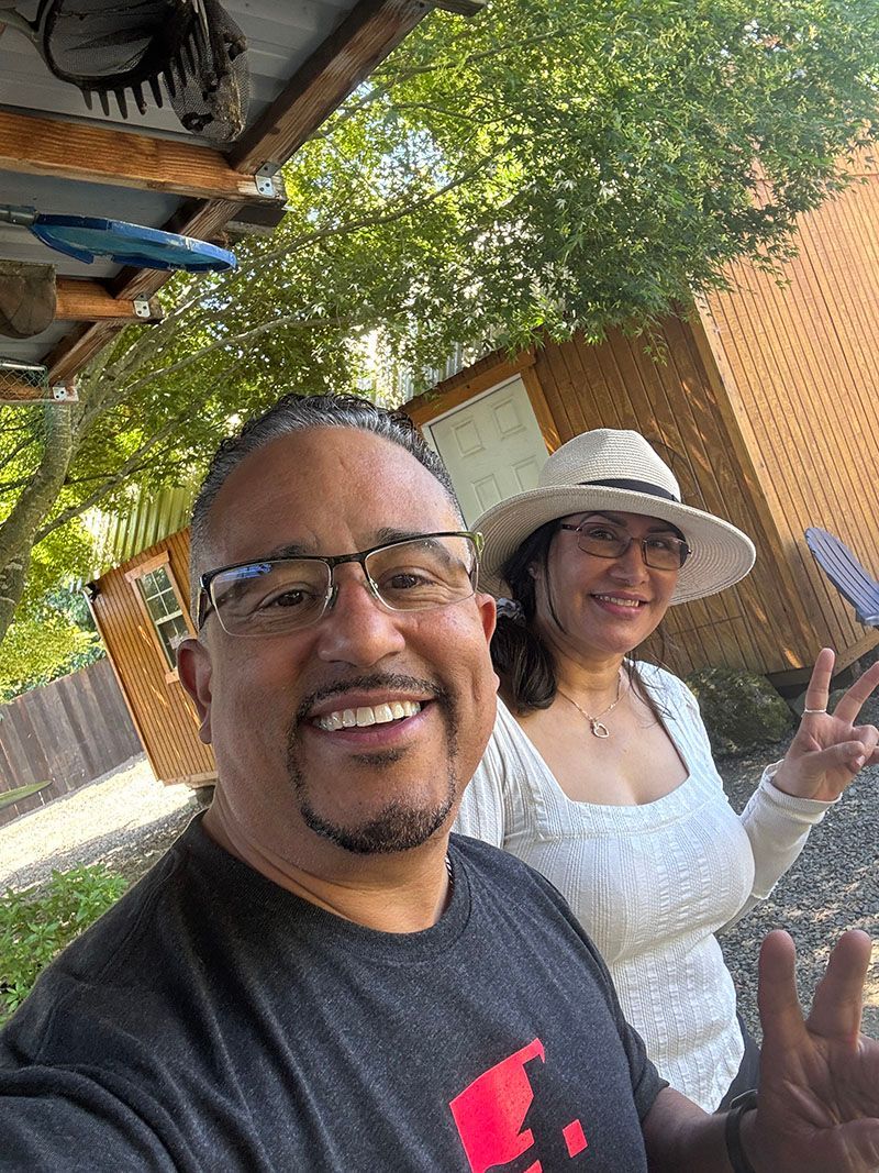 Man smiling with woman behind, both outdoors. Woman wearing hat and making peace sign.