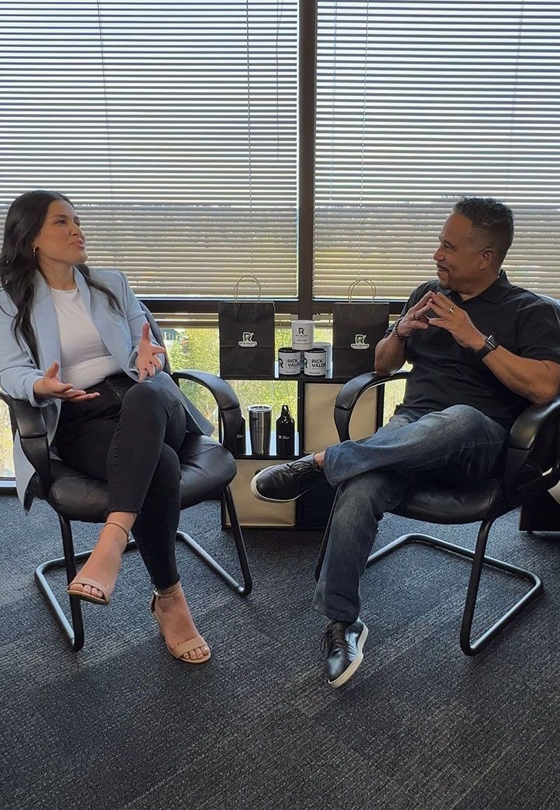 Two people seated, conversing. Woman in blazer, man in black shirt, both in chairs by window.