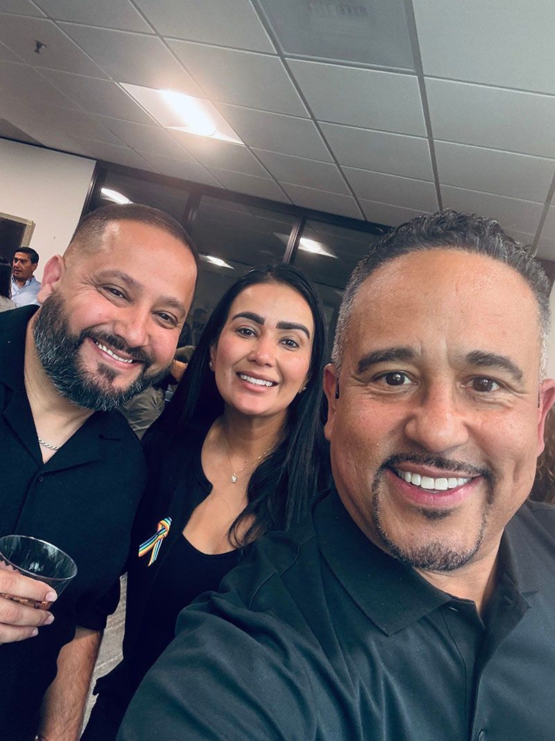 Three smiling people posing for a selfie indoors. The woman is in the middle, and two men flank her. All are dressed in black.