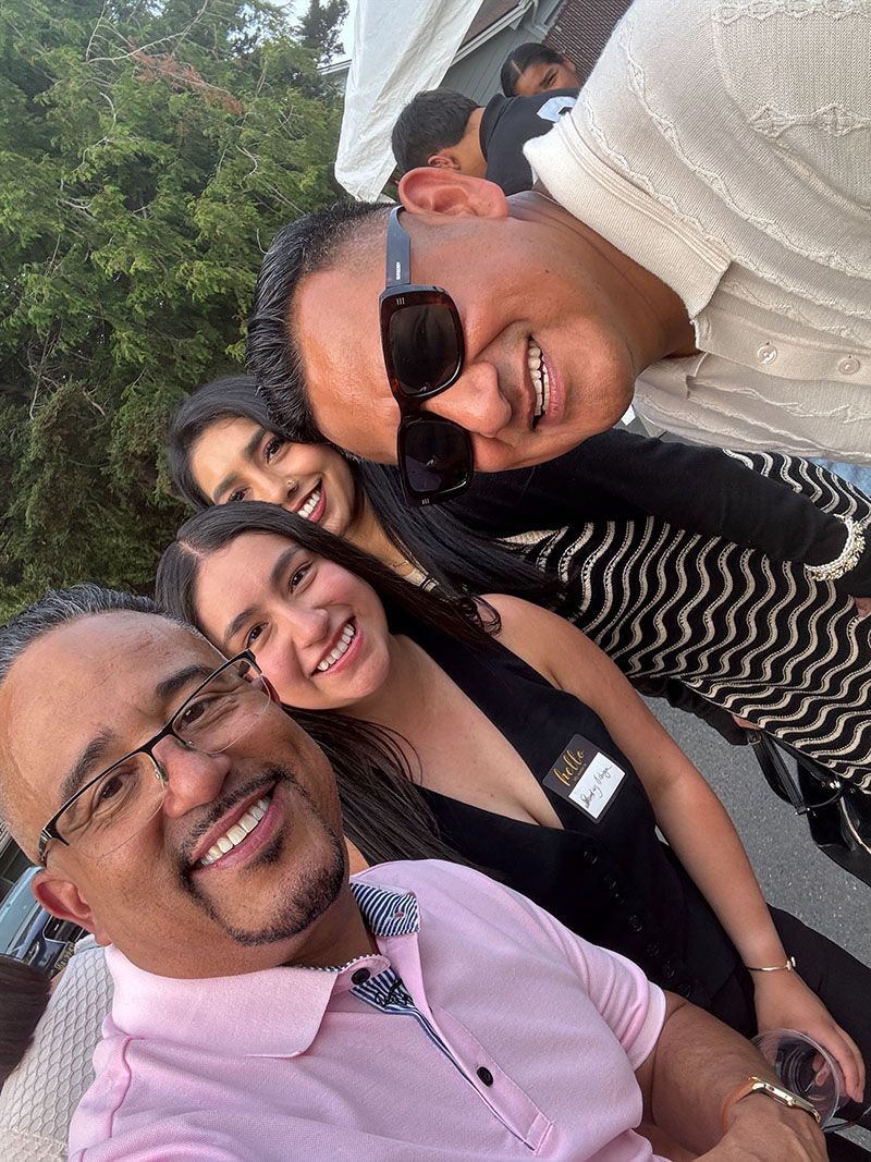 Group selfie: Smiling people outside. Man in pink shirt, woman in black, another woman with long dark hair.