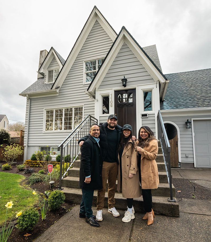 Four people smiling in front of a gray house with a peaked roof, steps, and a green lawn.