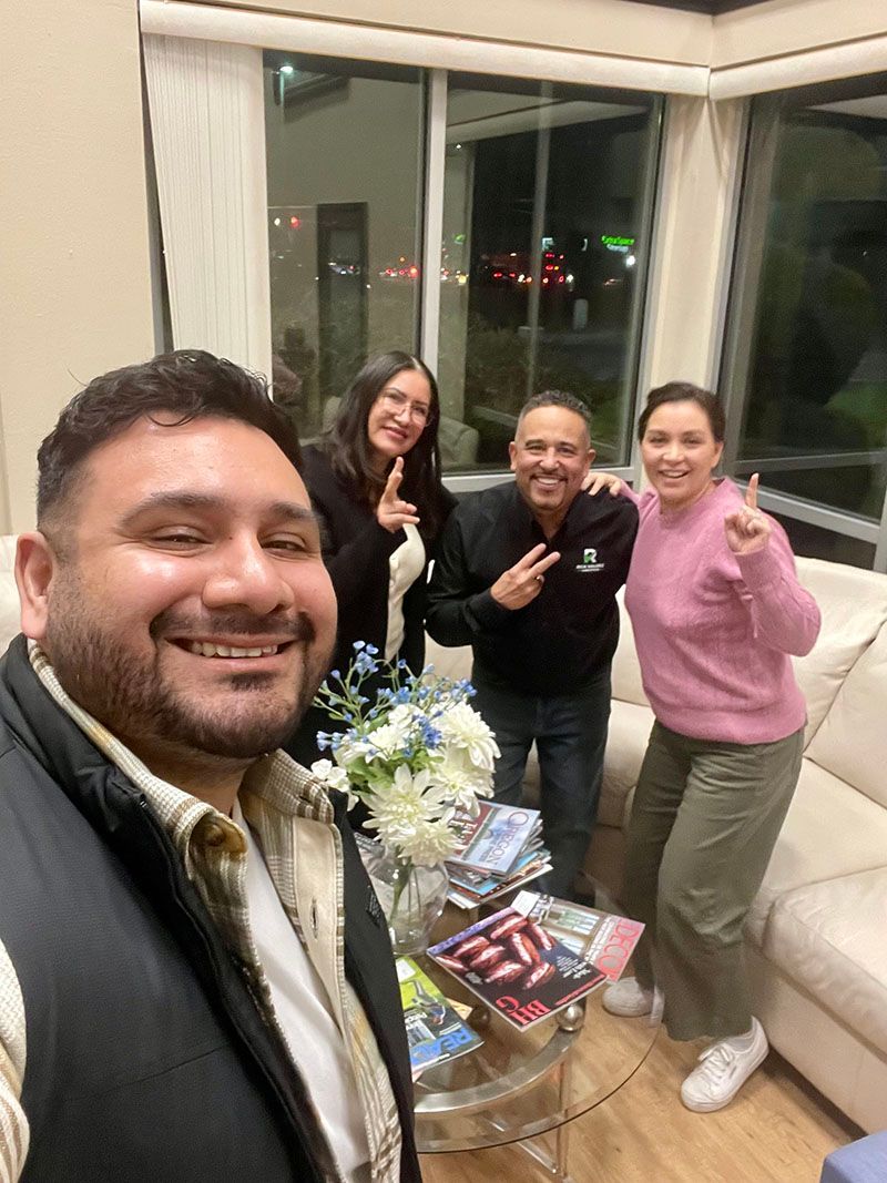 Four smiling people pose indoors, near a window and a sofa. They gesture, with books on a table.