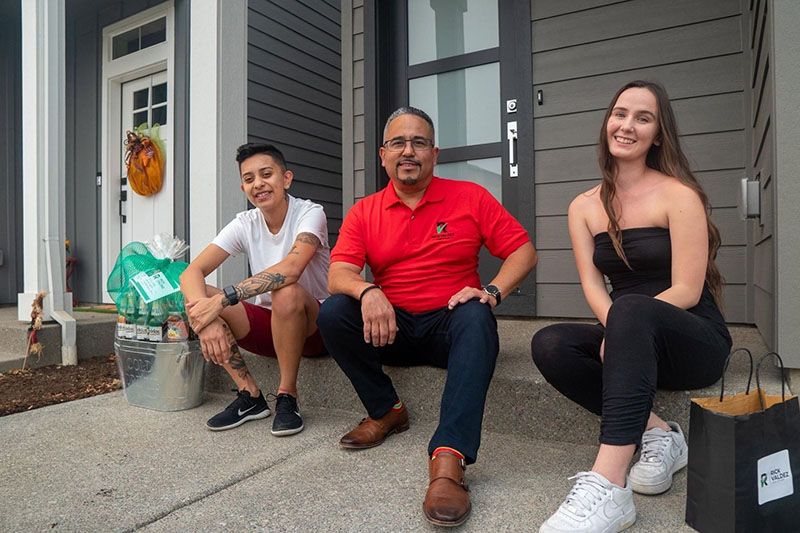 Three people sit on steps outside a house. Man in red shirt, two others. One with arm tattoos, one with a black outfit and bag.