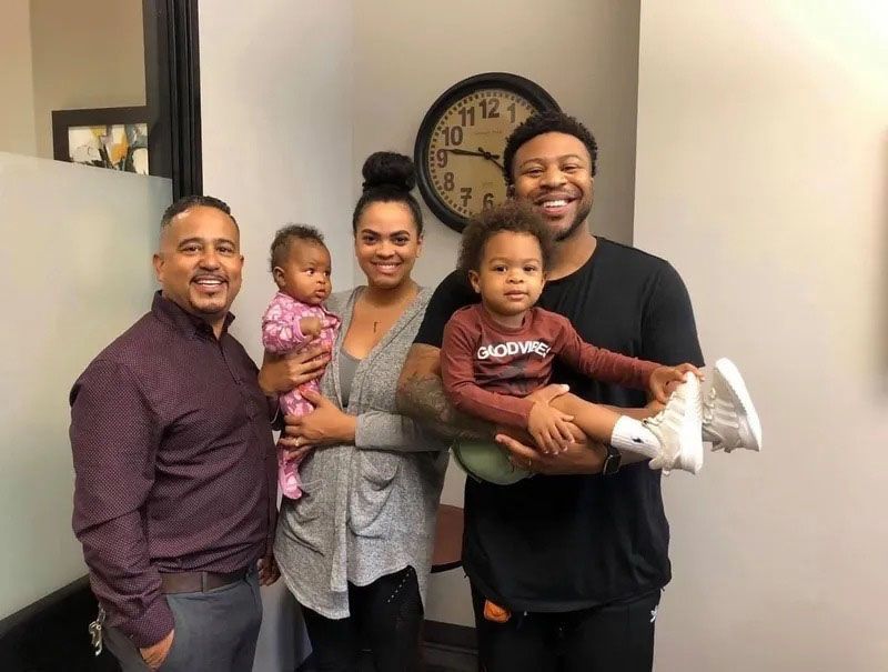 Family posing with a man, possibly in an office. They smile, one person holds a baby, another a toddler. A clock is on the wall.