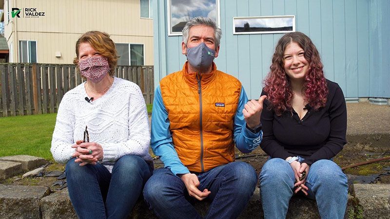 Three masked people sitting outside, smiling. The man in the center points to the right.
