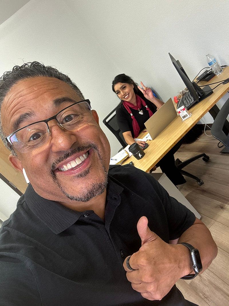 Man smiling, giving thumbs up, with a woman at a desk in the background. Office setting.