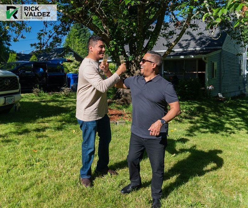 Two men bumping fists in a yard, house in background. One wears jeans, other black pants, both smiling.