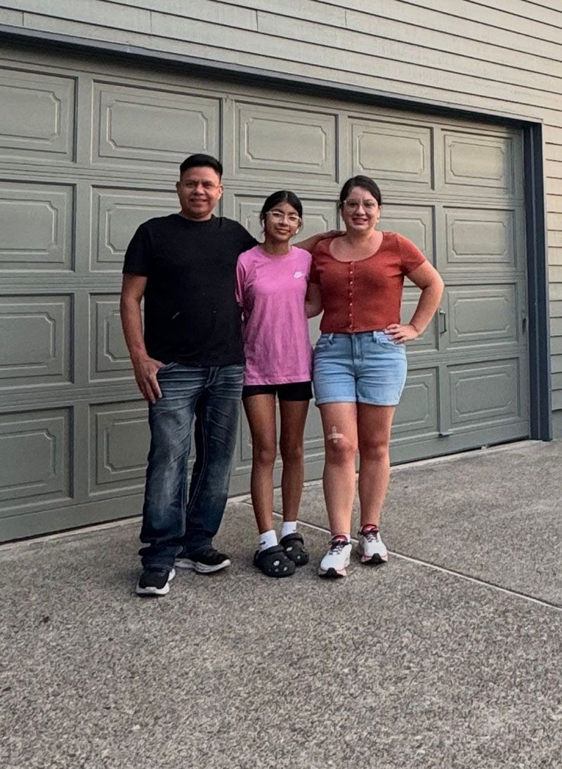 Three people pose in front of a gray garage door. Man in black shirt and jeans, girl in pink, and woman in shorts.