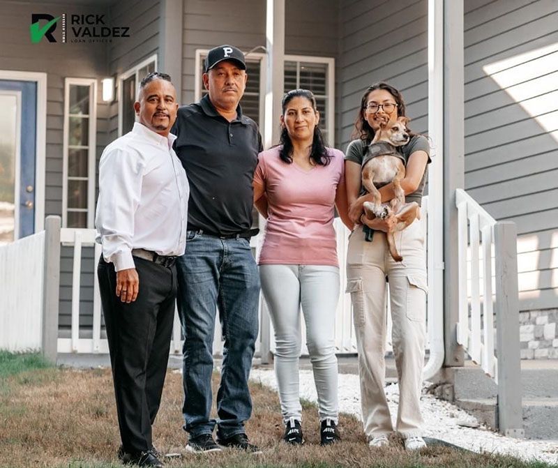 Group of four people standing outside a house. Woman holding a dog, others smiling.