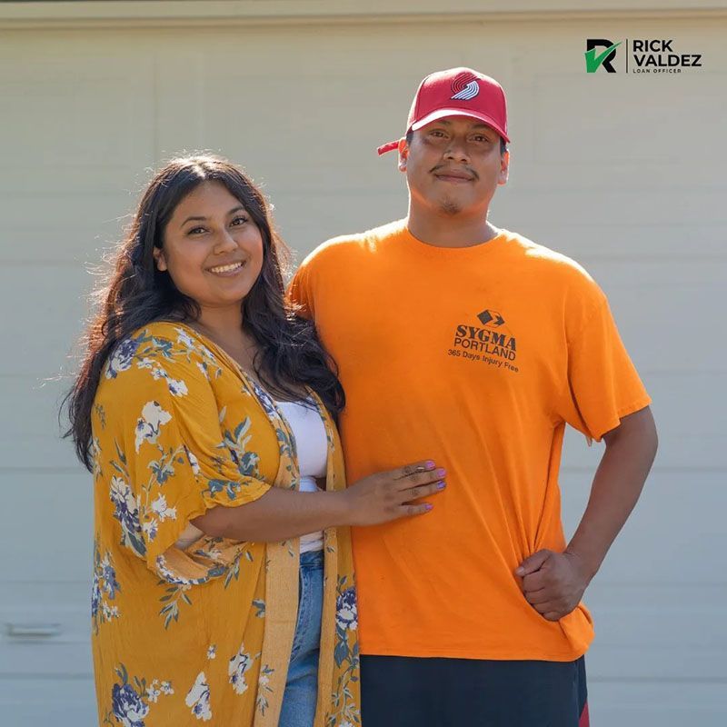 Smiling couple poses in front of a garage door. Woman in yellow floral top, man in orange shirt and baseball cap.