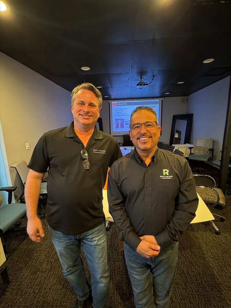 Two men smiling, posing together in a room with a projector screen. One in a black shirt and jeans, the other in a button-down shirt.