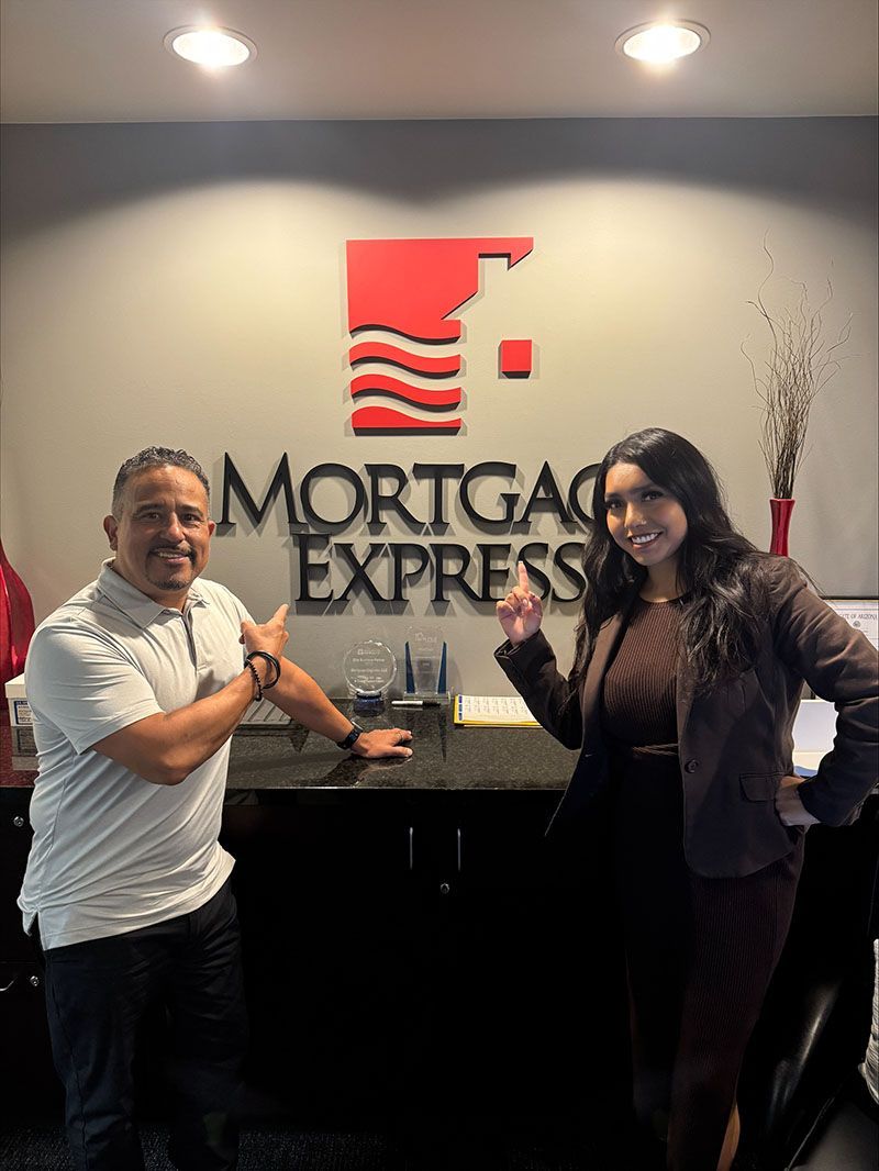 Two people pose by the Mortgage Express sign, giving thumbs up. Office setting with red and black logo.