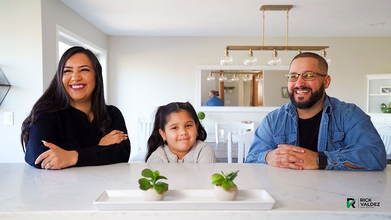 A family smiles together at a kitchen counter. Two small potted plants in the foreground.