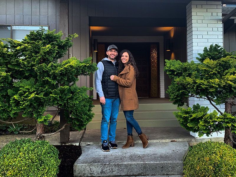 Couple stands on steps in front of a house; woman in brown coat, man in vest smiles.
