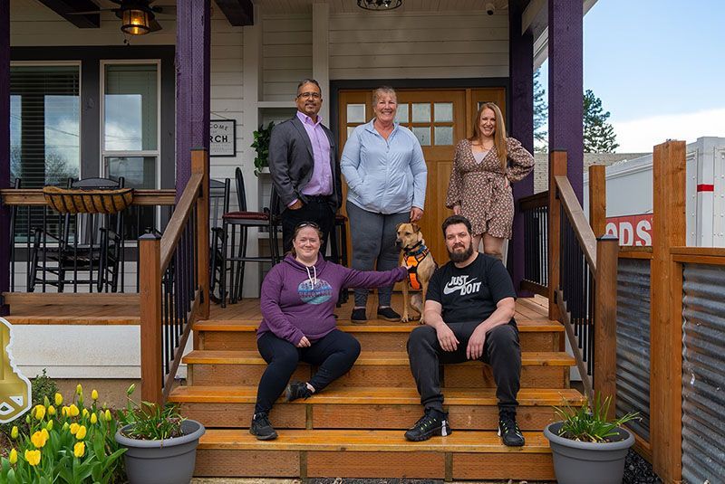 Group of people on porch steps, posing with a dog. Building has purple accents and outdoor seating.