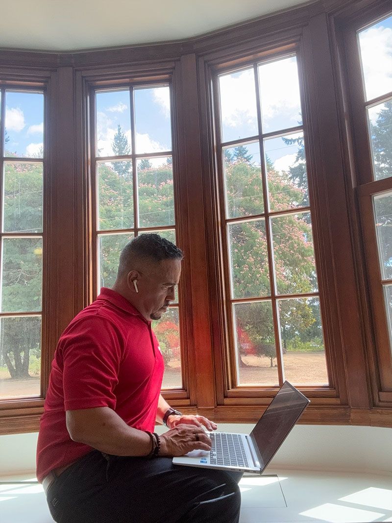 Man in red shirt working on a laptop by a window overlooking a lush garden.