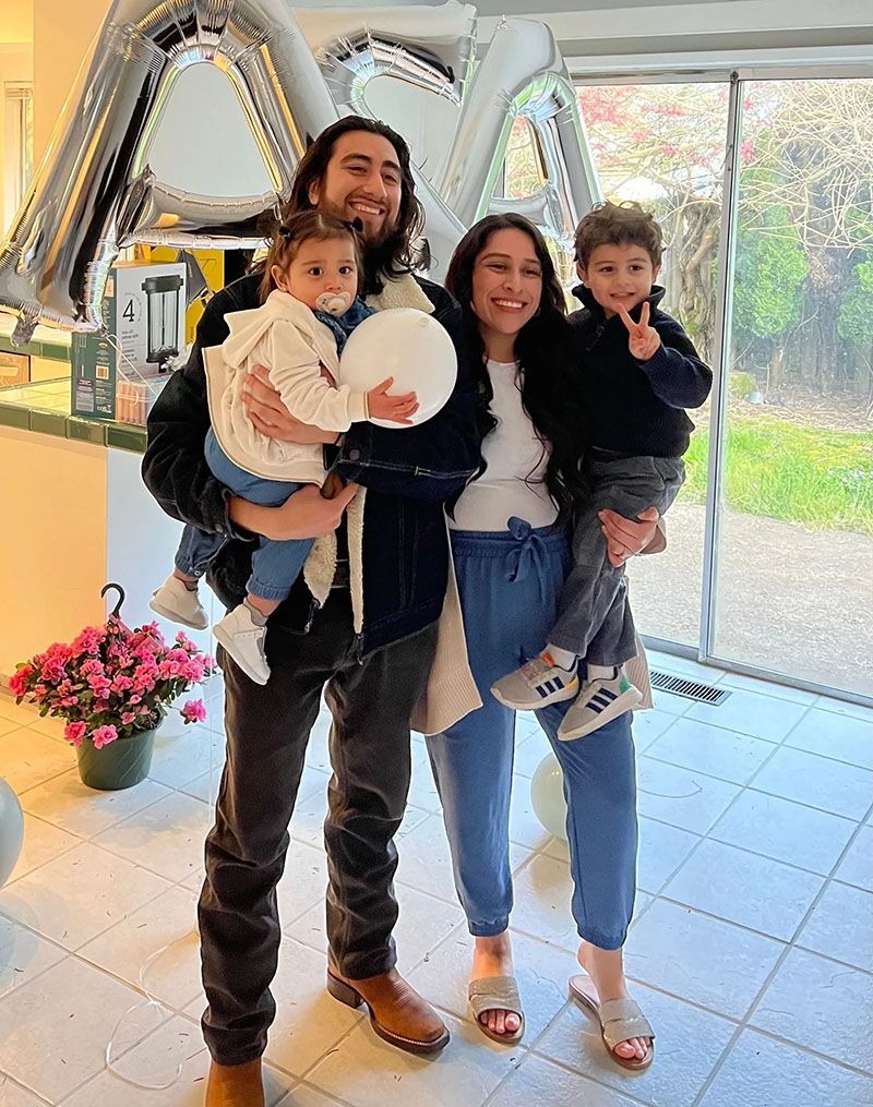 Family of four smiling, posing indoors near a window. Man holding baby, woman holding a round item, and another child. Balloons visible.