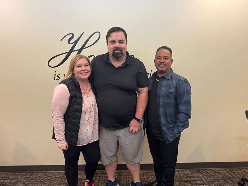 Three people posing for photo in front of wall with cursive text. Woman in vest smiles; two men look toward the camera.