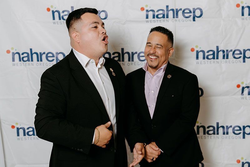 Two men in suits laugh together in front of a backdrop with logos.